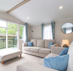 A living room with two beige sofas blue cushions a beige ottoman a round mirror and a window with blue curtains at Eltons Escape in Pony Meadow 3 White Cross Bay near Windermere