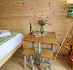 A wooden room with a bed, table with flowers, wine bottle, glasses, stools, and a floor lamp at Moel Arthur in Bryniau Clwyd near Denbigh