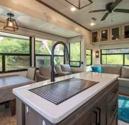 An interior view of an RV kitchen and living area with a kitchen island featuring a sink and faucet a dining table with chairs beige sofas with blue pillows and large windows at Tickeydoo Tor Down Quarry in St Breward