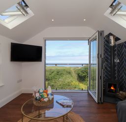 A living room with a glass coffee table basket with groceries a wall-mounted television a fireplace with black tiles and an open door showing a green landscape and sea view at Victory View in Pendeen