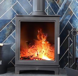 A wood burning stove with fire inside a room with blue tiled walls and a black ash bucket and fireplace tools nearby at Victory View Pendeen