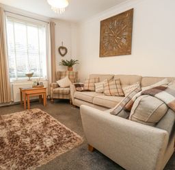 A living room with a beige sofa and armchair a wooden side table underneath a window with curtains and a wall clock at Garibaldi Cottage in Weymouth
