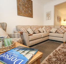 A living room with a beige corner sofa and an armchair with cushions a wooden table with plants and a book and a clock on the wall at Garibaldi Cottage in Weymouth