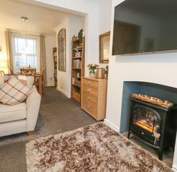 A living room with a sofa and cushions a television above a fireplace and a dining area with a table and chairs at Garibaldi Cottage in Weymouth