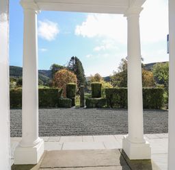 View from a doorway with white columns onto a gravel pathway with trimmed hedges and trees in the background at Llwynderw Hall in Abergwesyn near Llanwrtyd Wells
