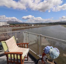 A balcony with a green chair and a table with flowers and drinks overlooking a river and a train on a track at Estuary Escape in Porthmadog