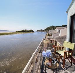 A balcony with a chair small table with flowers and a wine bottle overlooking a river and houses along the shore at Estuary Escape in Porthmadog