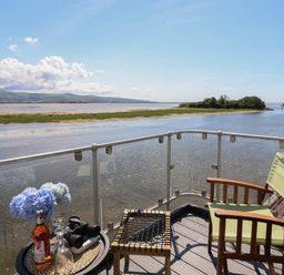 A balcony with a chair a small table holding a bottle flowers glasses and binoculars overlooking a river and landscape at Estuary Escape in Porthmadog