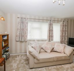 A living room with a beige sofa and floral cushions a wooden bookshelf a television and patterned curtains at Ground Floor Annex in Milton-Under-Wychwood