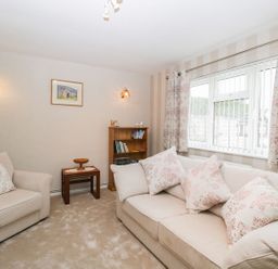 A living room with beige sofas and floral cushions a wooden bookshelf and side tables at Ground Floor Annex in Milton-Under-Wychwood