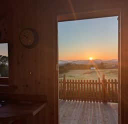 A wooden kitchen interior with a clock on the wall and an open door leading to a deck overlooking a sunrise and fields at Hafan Gelli Sheperds Hut - Tudwal in Pwllheli