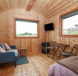 A wooden interior room with a blue sofa and pillows a small round table a wall-mounted television a small dining table with two chairs and a bed Hafan Gelli Sheperds Hut - Tudwal Pwllheli