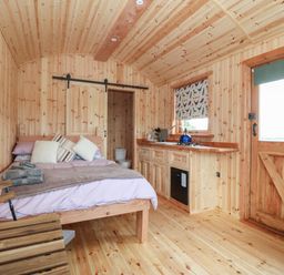 An interior of a wooden room with a bed, kitchen area, wooden door, and a sliding barn door at Hafan Gelli Sheperds Hut - Tudwal in Pwllheli