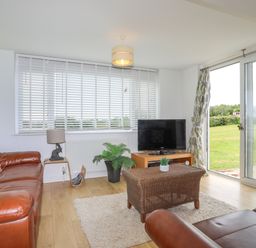 A living room with brown leather sofas a TV on a wooden stand and sliding glass doors opening to a garden at Mor Glas in Abersoch