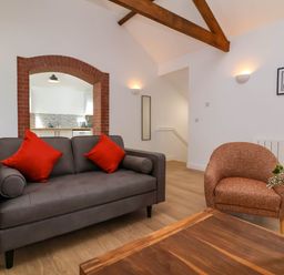 A living room with a gray sofa with red cushions an orange armchair a wooden coffee table and a view into a kitchen through a brick arch at Woodland Roost Parkham near Bideford