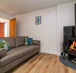 A living room with a brown sofa and two green leaf patterned cushions a wood burning stove with firewood and a small framed picture on the wall at Ladybird Rest in Parkham near Bideford