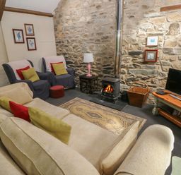 A living room with a stone wall and wood burning stove with beige sofa armchairs and a television at The Old Stable in Pontfaen near Fishguard