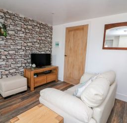 A living room with a stone patterned wallpaper wall a wood tv stand with a television a beige ottoman a cream sofa and a mirror on the wall at Field View in Llanybydder