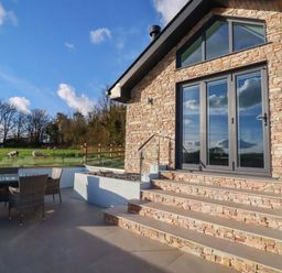 An outdoor patio area with a stone house and glass doors next to a table with chairs and a grassy field with sheep at Lake View in Kestle Mill near Quintrell Downs