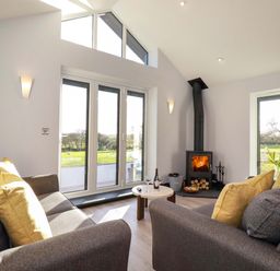 A living room with gray sofas and yellow pillows a round wooden table a wood burning stove and large windows at Lake View in Kestle Mill near Quintrell Downs