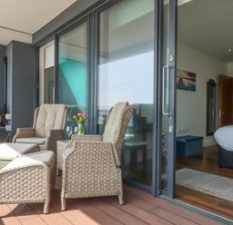 A balcony with wicker chairs and ottomans next to a sliding glass door leading to a bedroom at Apartment 17 Abbey Sands in Torquay