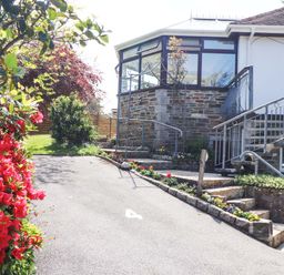 Driveway with flowers along the edges and stone steps leading to a house with large windows and a clock on the wall at Lake View Swanpool Nr Falmouth