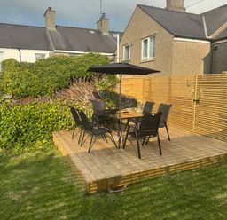 A wooden deck with a table umbrella and six chairs in a garden surrounded by greenery and houses at 1 Mona Terrace in Llanfachraeth