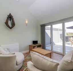 A living room with two beige armchairs a wooden coffee table a small TV on a wooden stand a patterned rug and sliding glass doors to a patio at Singletree Cottage in Swainsford near Mere