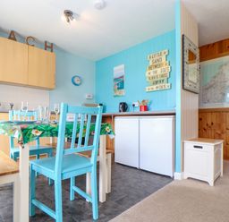 A kitchen and dining area with blue chairs a table with a floral tablecloth wooden cabinets and a sign on the wall at Skylarks Trevellas Nr St Agnes