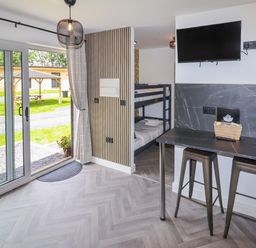 A room with bunk beds a wall-mounted television a counter with two stools and sliding glass doors overlooking a grassy area at Valley Lodge 4 - North Wales Eco Lodges in Graigfechan near Ruthin