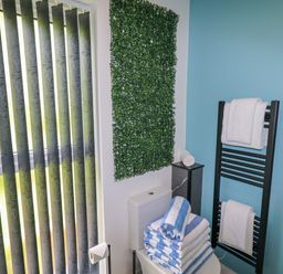 A bathroom with vertical blinds a toilet with stacked blue and white towels a towel rack with white towels and a green wall decoration at Valley Lodge 4 - North Wales Eco Lodges in Graigfechan near Ruthin