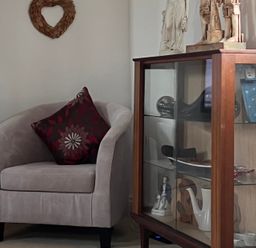 A beige armchair with a patterned cushion next to a wooden glass display cabinet with various decorative items at 20 Keay Heights in St Austell