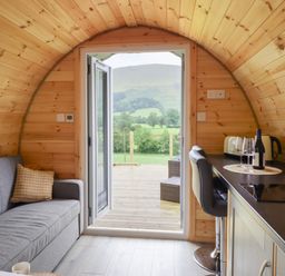 A small wooden cabin interior with a gray sofa, kitchenette with bar stools, and open double doors leading to a deck with a view of green hills at Caban Tirion in Dinas Mawddwy near Dolgellau
