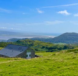 A stone house with a dark roof on a green hillside with hills and a body of water in the background at Beudy Mawr in Cwmystradllyn near Tremadog