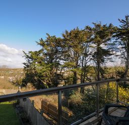 A balcony with a chair overlooking trees and a fenced yard at Surf Break in Polzeath