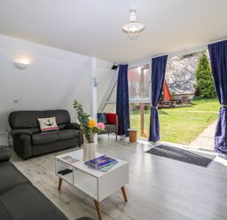 A living room with black sofas a white coffee table with magazines and flowers and large glass doors with blue curtains overlooking a garden at Chalet 58 - The Beach House in Kingsdown