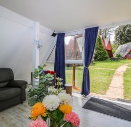 A living room with a black sofa and flowers on a table next to a glass door with blue curtains showing a garden outside at Chalet 58 - The Beach House in Kingsdown