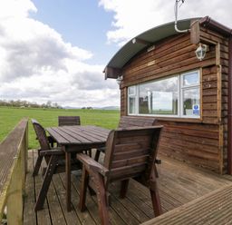 An outdoor wooden deck with a table and four chairs next to a wooden cabin in a grassy field at Hollie in Raglan