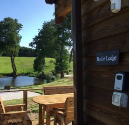 Wooden chairs and table on a deck near a pond with green trees and grass at Hollie Lodge Raglan