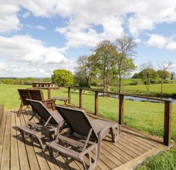 A wooden deck with lounge chairs and a table overlooking a grassy field and a river at Poppy in Raglan