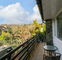 A small balcony with a glass round table and two chairs overlooking trees and neighboring houses at Lyndhurst in Ambleside