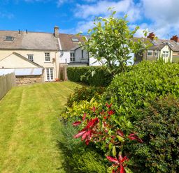 A backyard garden with green grass, bushes, and trees behind houses at Orielton Cottage in Dinas Cross