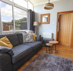 A living room with a gray sofa with cushions a floor lamp wooden nested tables and a large window at Pebble Beach Chalet in Seaton