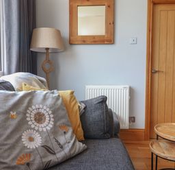 A living room corner with a grey sofa with cushions a floor lamp a wooden framed mirror a radiator a wooden door and nesting wooden tables at Pebble Beach Chalet in Seaton