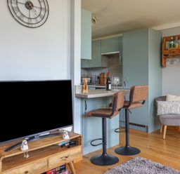 A living area with a television on a wooden stand a kitchen counter with two bar stools and a gray chair with cushions at Pebble Beach Chalet in Seaton