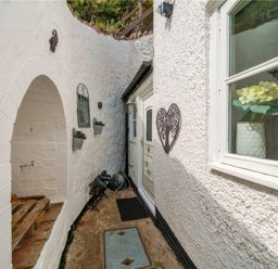 A narrow outdoor passageway with white textured walls a white door a window with a vase of flowers and decorative metal wall hangings at Holly Cottage in Great Malvern