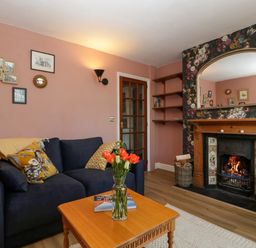 A living room with a navy sofa, wooden coffee table with flowers, fireplace with mirror and floral wallpaper at Holly Cottage in Great Malvern
