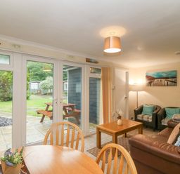 A living room with a brown sofa two armchairs wooden coffee and dining tables and glass doors opening to a garden at 245 Riverside Lodge in Caeathro
