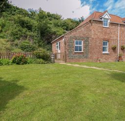 A brick house with tiled roof and white framed windows beside a garden with grass flowers and bushes at Dairy Cottage in West Caister near Caister-on-Sea