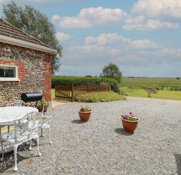An outdoor patio area with white metal table and chairs gravel ground two flower pots stone wall building and open field at Flint Cottage in West Caister near Caister-on-Sea